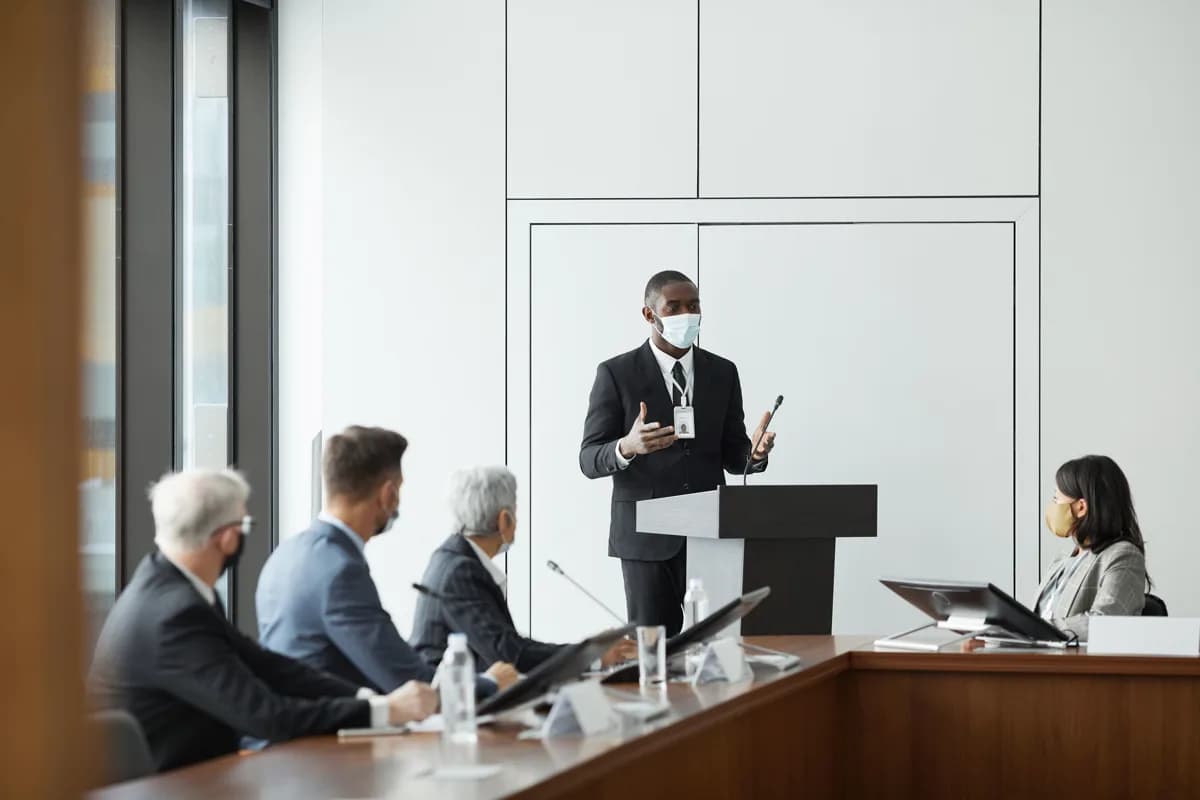 A presenter addressing a panel of masked colleagues in a conference room during a compliance briefing